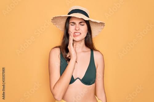 Young beautiful girl wearing swimwear bikini and summer sun hat over yellow background touching mouth with hand with painful expression because of toothache or dental illness on teeth. Dentist