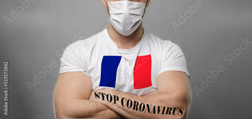 A young strong man in a white T-shirt with the image of the flag France crossed his arms over his chest stands on a light backgrou facing the camera. On hand tattoo Stop Coronavirus. Close-up.