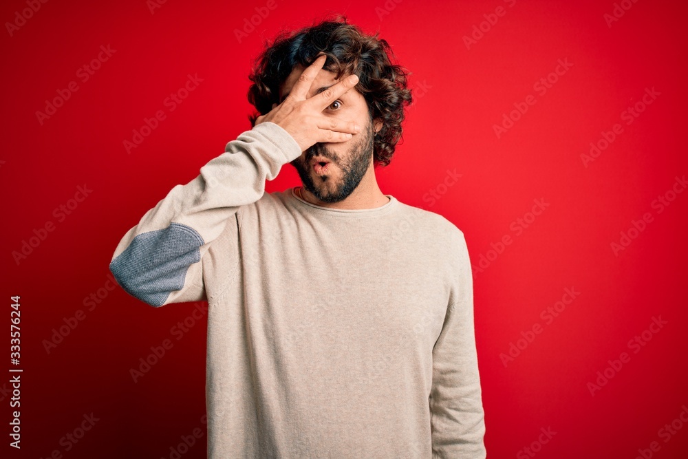 Young handsome man with beard wearing casual sweater standing over red background peeking in shock covering face and eyes with hand, looking through fingers with embarrassed expression. Young handsome man with beard wearing casual sweater standing over red background peeking in shock covering face and eyes with hand, looking through fingers with embarrassed expression.