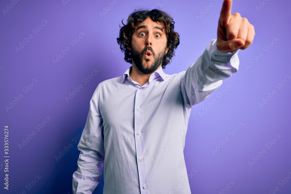 Young handsome business man with beard wearing shirt standing over purple background Pointing with finger surprised ahead, open mouth amazed expression, something on the front