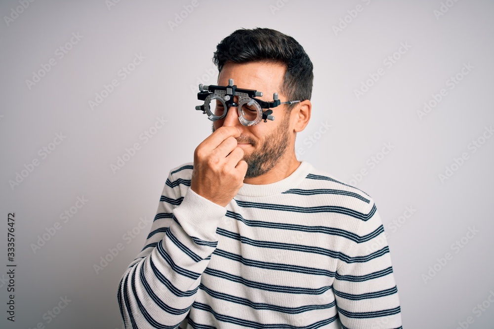 Young handsome man with beard wearing optometry glasses over isolated white background smelling something stinky and disgusting, intolerable smell, holding breath with fingers on nose. Bad smell Young handsome man with beard wearing optometry glasses over isolated white background smelling something stinky and disgusting, intolerable smell, holding breath with fingers on nose. Bad smell