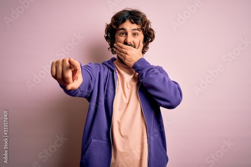 Young handsome sporty man with beard wearing casual sweatshirt over pink background laughing at you  pointing finger to the camera with hand over mouth  shame expression