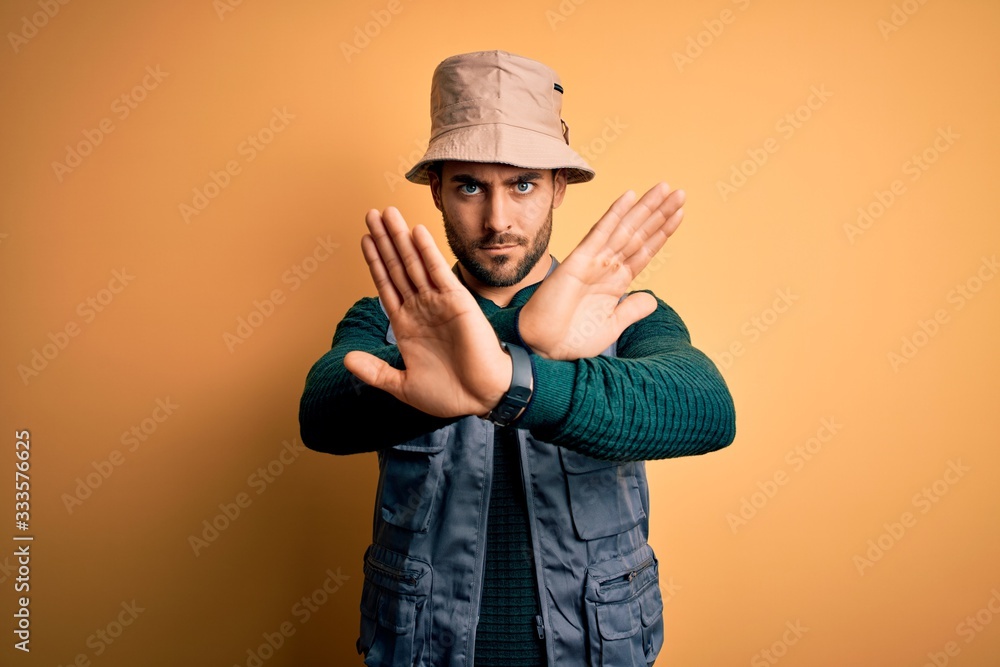 Handsome tourist man with beard on vacation wearing explorer hat over yellow background Rejection expression crossing arms and palms doing negative sign, angry face Handsome tourist man with beard on vacation wearing explorer hat over yellow background Rejection expression crossing arms and palms doing negative sign, angry face