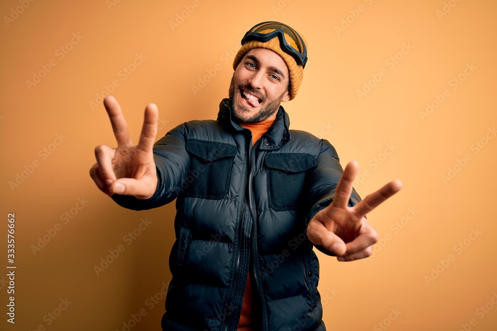 Young handsome skier man with beard wearing snow sportswear and ski goggles smiling with tongue out showing fingers of both hands doing victory sign. Number two. Young handsome skier man with beard wearing snow sportswear and ski goggles smiling with tongue out showing fingers of both hands doing victory sign. Number two.