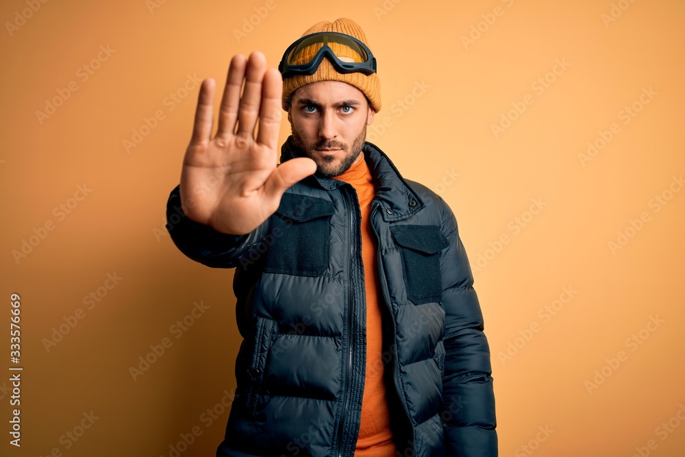 Young handsome skier man with beard wearing snow sportswear and ski goggles doing stop sing with palm of the hand. Warning expression with negative and serious gesture on the face. Young handsome skier man with beard wearing snow sportswear and ski goggles doing stop sing with palm of the hand. Warning expression with negative and serious gesture on the face.