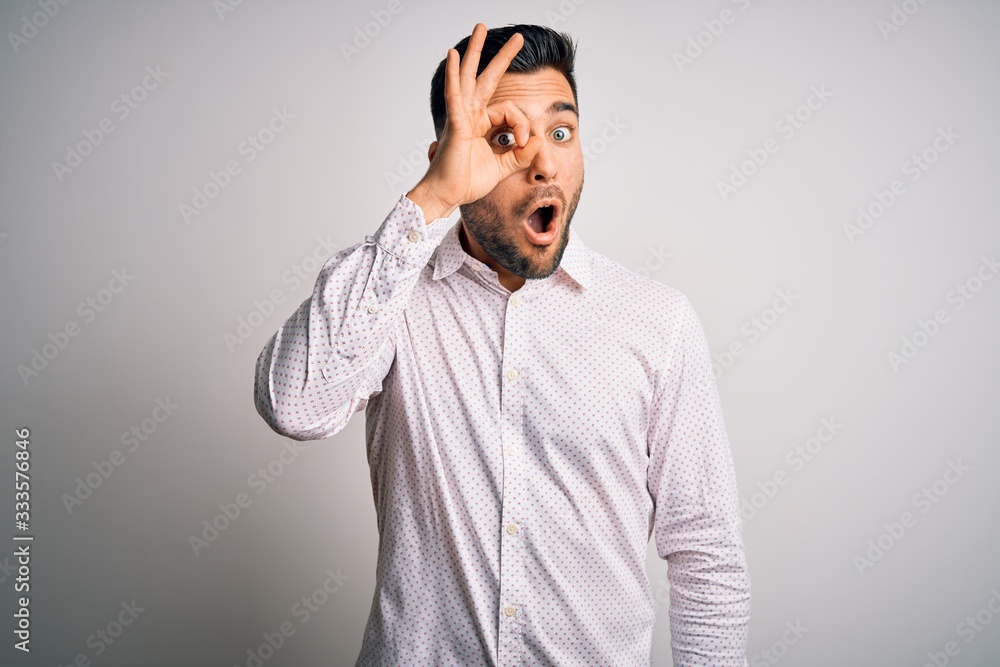 Young handsome man wearing elegant shirt standing over isolated white background doing ok gesture shocked with surprised face, eye looking through fingers. Unbelieving expression.