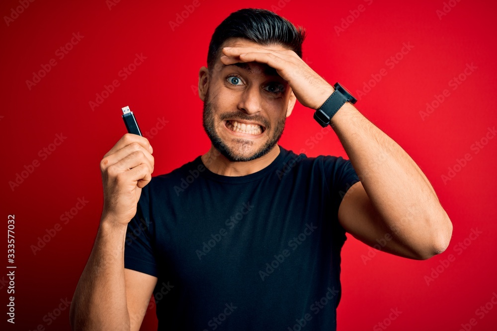 Young man holding USB memory data drive over red isolated background stressed with hand on head, shocked with shame and surprise face, angry and frustrated. Fear and upset for mistake. Young man holding USB memory data drive over red isolated background stressed with hand on head, shocked with shame and surprise face, angry and frustrated. Fear and upset for mistake.