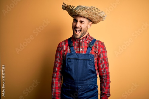 Young rural farmer man wearing bib overall and countryside hat over yellow background winking looking at the camera with sexy expression, cheerful and happy face.