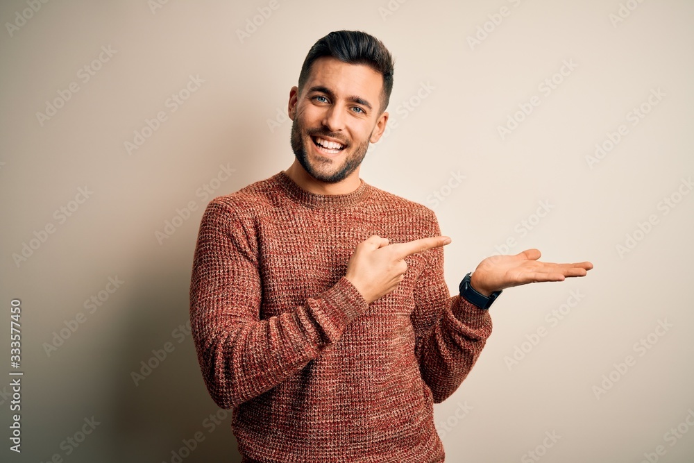 Young handsome man wearing casual sweater standing over isolated white background amazed and smiling to the camera while presenting with hand and pointing with finger. Young handsome man wearing casual sweater standing over isolated white background amazed and smiling to the camera while presenting with hand and pointing with finger.