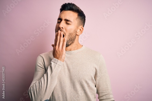 Young handsome man wearing casual sweater standing over isolated pink background bored yawning tired covering mouth with hand. Restless and sleepiness.