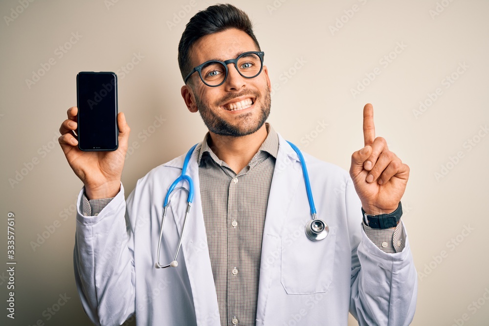 Young doctor man wearing stethoscope showing smartphone screen over isolated background surprised with an idea or question pointing finger with happy face, number one Young doctor man wearing stethoscope showing smartphone screen over isolated background surprised with an idea or question pointing finger with happy face, number one