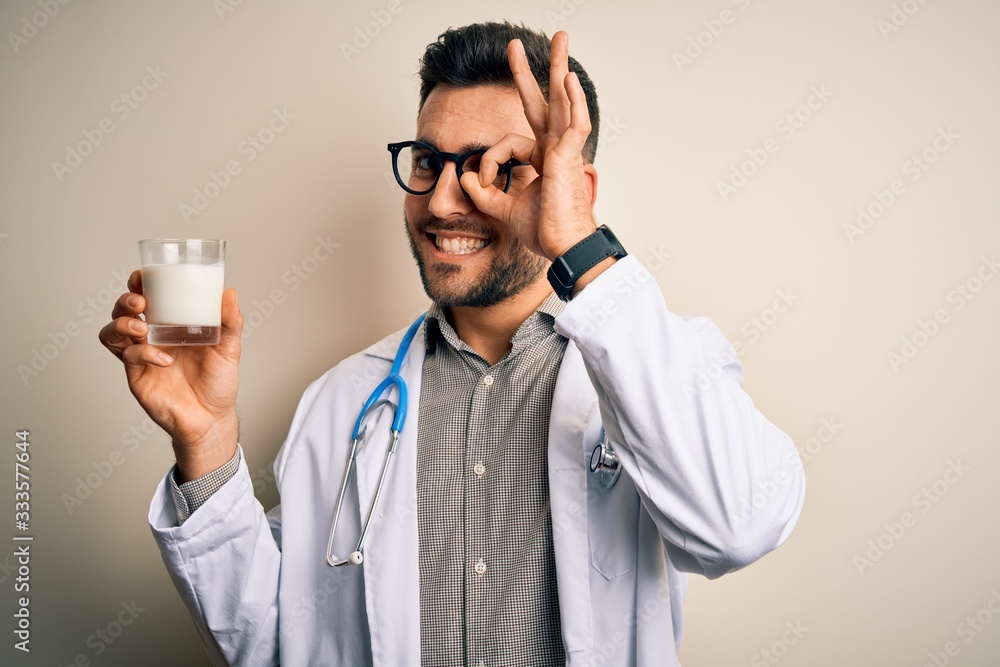 Young doctor man wearing stethoscope holding a glass of milk over isolated background with happy face smiling doing ok sign with hand on eye looking through fingers Young doctor man wearing stethoscope holding a glass of milk over isolated background with happy face smiling doing ok sign with hand on eye looking through fingers
