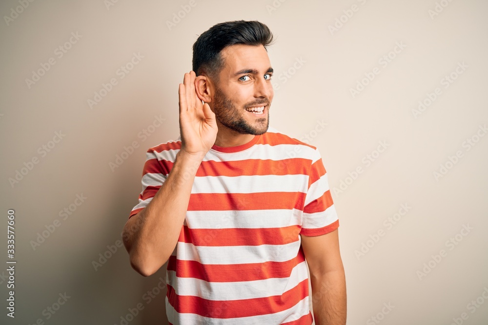 Young handsome man wearing casual striped t-shirt standing over isolated white background smiling with hand over ear listening an hearing to rumor or gossip. Deafness concept. Young handsome man wearing casual striped t-shirt standing over isolated white background smiling with hand over ear listening an hearing to rumor or gossip. Deafness concept.