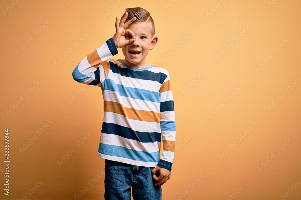 Young little caucasian kid with blue eyes wearing colorful striped shirt over yellow background doing ok gesture with hand smiling, eye looking through fingers with happy face. Young little caucasian kid with blue eyes wearing colorful striped shirt over yellow background doing ok gesture with hand smiling, eye looking through fingers with happy face.