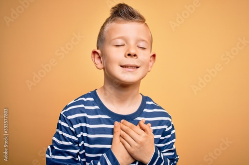 Young little caucasian kid with blue eyes wearing nautical striped shirt over yellow background smiling with hands on chest with closed eyes and grateful gesture on face. Health concept.