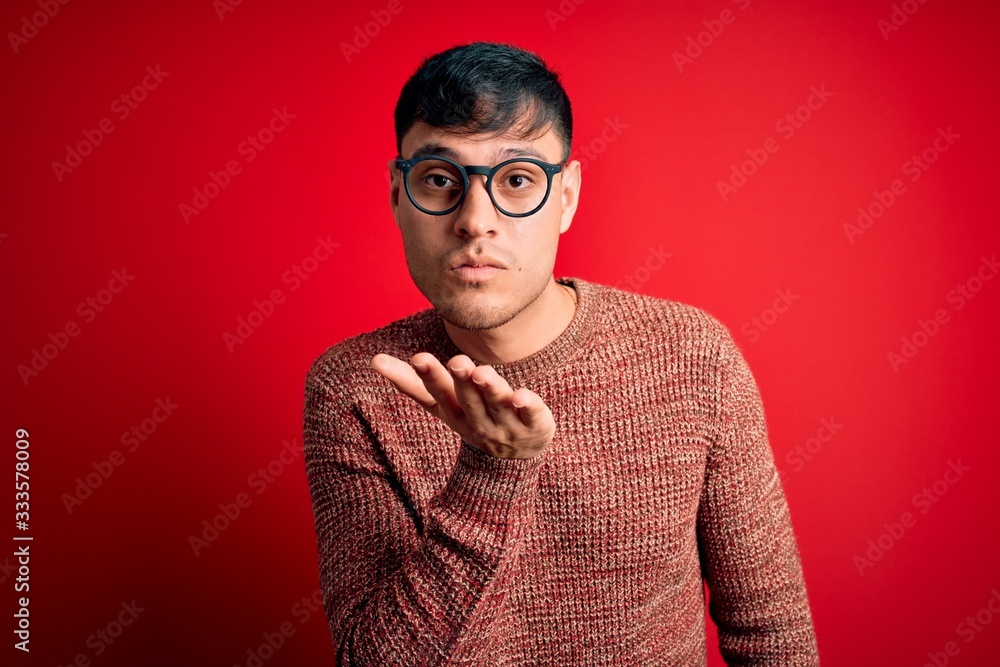Young handsome hispanic man wearing nerd glasses over red background looking at the camera blowing a kiss with hand on air being lovely and sexy. Love expression. Young handsome hispanic man wearing nerd glasses over red background looking at the camera blowing a kiss with hand on air being lovely and sexy. Love expression.
