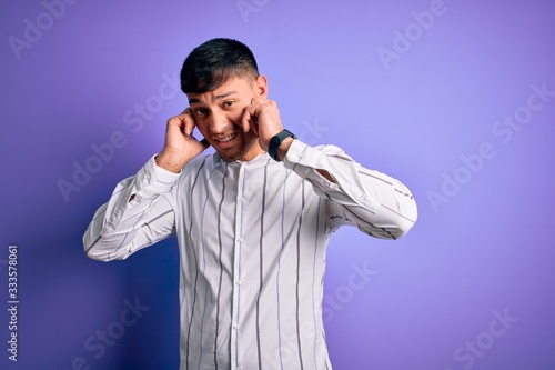 Young handsome hispanic man wearing elegant business shirt standing over purple background covering ears with fingers with annoyed expression for the noise of loud music. Deaf concept.