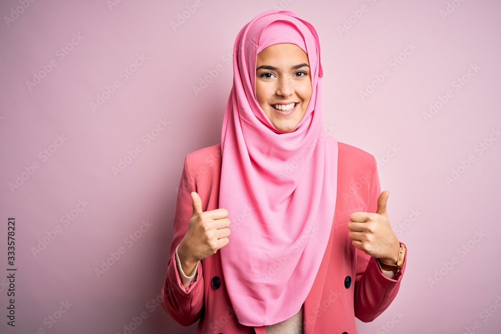 Young beautiful girl wearing muslim hijab standing over isolated pink background success sign doing positive gesture with hand, thumbs up smiling and happy. Cheerful expression and winner gesture. Young beautiful girl wearing muslim hijab standing over isolated pink background success sign doing positive gesture with hand, thumbs up smiling and happy. Cheerful expression and winner gesture.