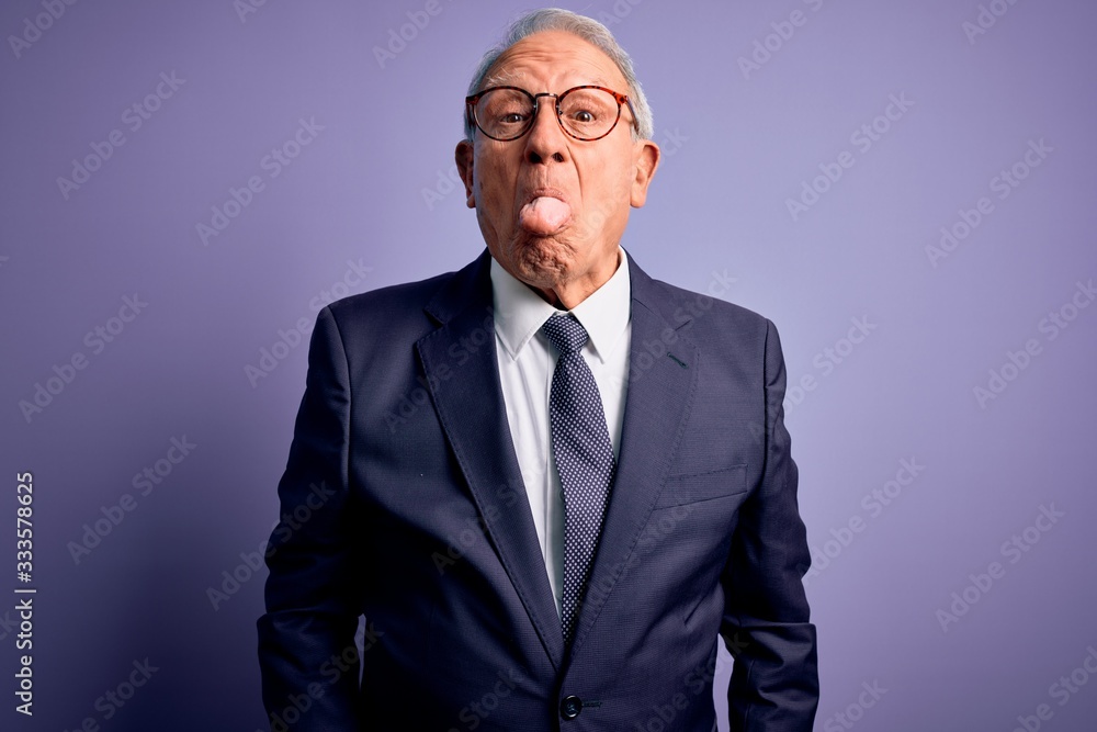 Grey haired senior business man wearing glasses and elegant suit and tie over purple background sticking tongue out happy with funny expression. Emotion concept. Grey haired senior business man wearing glasses and elegant suit and tie over purple background sticking tongue out happy with funny expression. Emotion concept.
