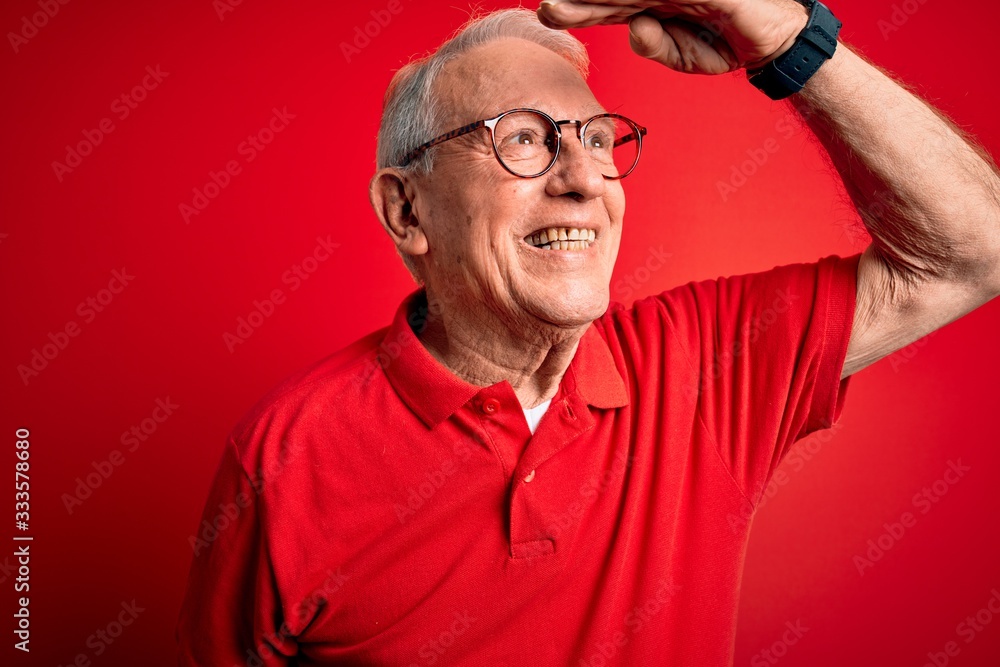 Grey haired senior man wearing glasses and casual t-shirt over red background very happy and smiling looking far away with hand over head. Searching concept.