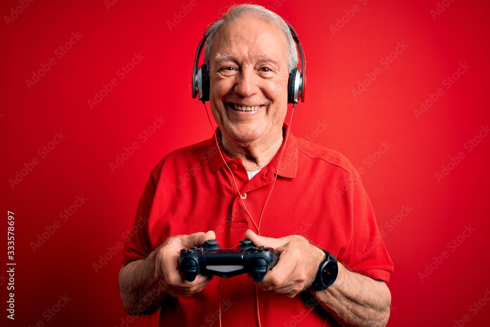 Senior grey haired gamer man playing video games using gamepad joystick over red background with a happy face standing and smiling with a confident smile showing teeth Senior grey haired gamer man playing video games using gamepad joystick over red background with a happy face standing and smiling with a confident smile showing teeth