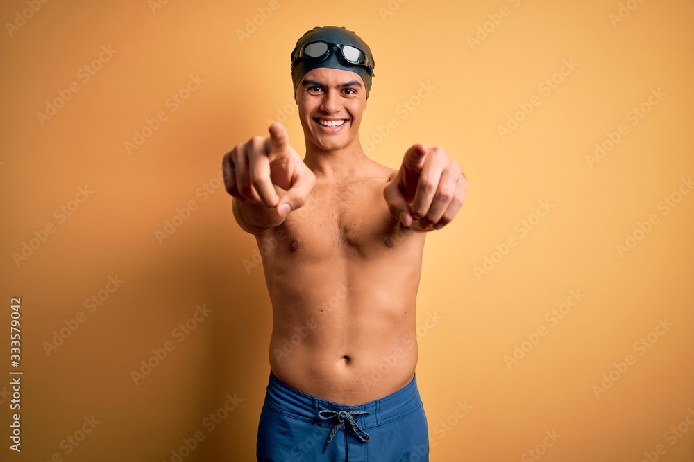 Young handsome man shirtless wearing swimsuit and swim cap over isolated yellow background pointing to you and the camera with fingers, smiling positive and cheerful