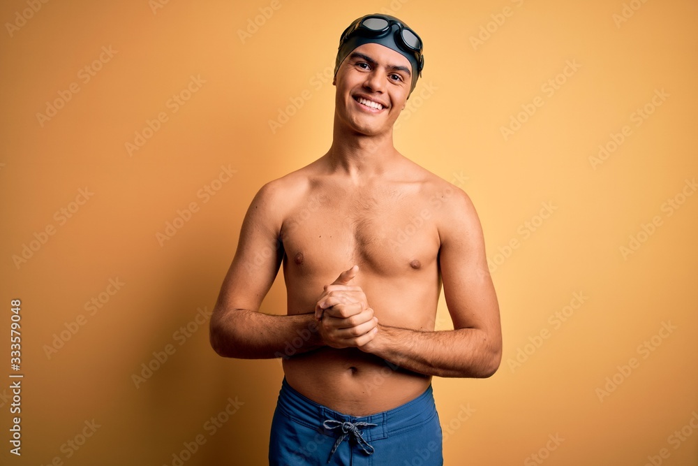 Young handsome man shirtless wearing swimsuit and swim cap over isolated yellow background with hands together and crossed fingers smiling relaxed and cheerful. Success and optimistic