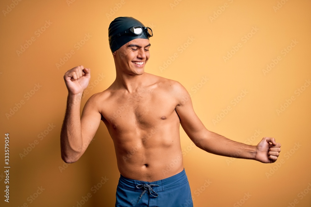 Young handsome man shirtless wearing swimsuit and swim cap over isolated yellow background Dancing happy and cheerful, smiling moving casual and confident listening to music