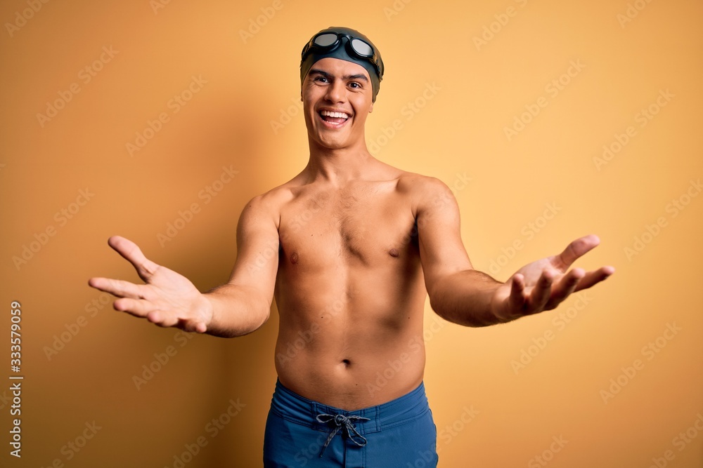 Young handsome man shirtless wearing swimsuit and swim cap over isolated yellow background smiling cheerful offering hands giving assistance and acceptance.