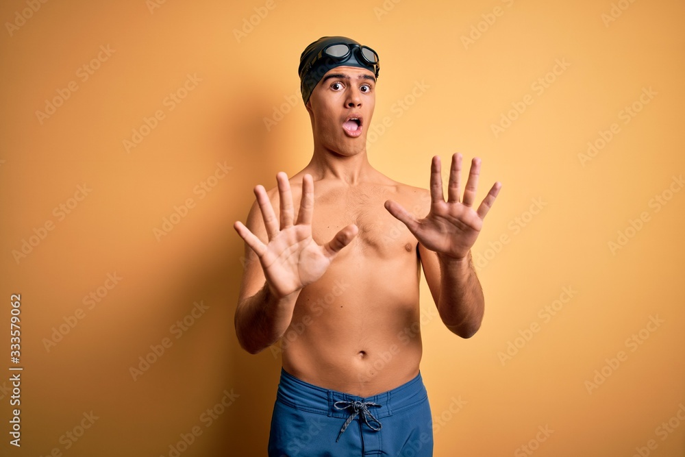 Young handsome man shirtless wearing swimsuit and swim cap over isolated yellow background afraid and terrified with fear expression stop gesture with hands, shouting in shock. Panic concept.