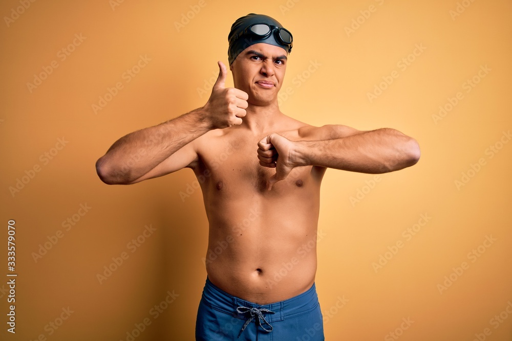 Young handsome man shirtless wearing swimsuit and swim cap over isolated yellow background Doing thumbs up and down, disagreement and agreement expression. Crazy conflict