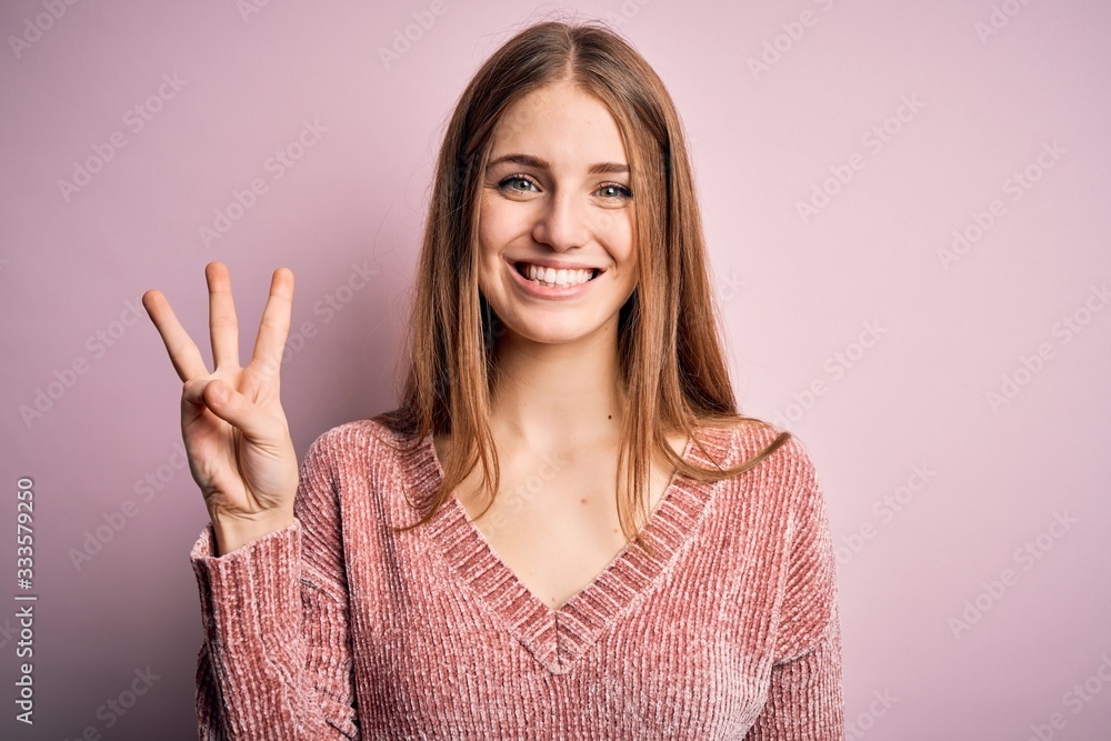 Young beautiful redhead woman wearing casual sweater over isolated pink background showing and pointing up with fingers number three while smiling confident and happy. - obrazy, fototapety, plakaty 