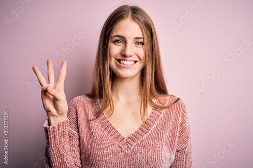 Young beautiful redhead woman wearing casual sweater over isolated pink background showing and pointing up with fingers number three while smiling confident and happy. photo