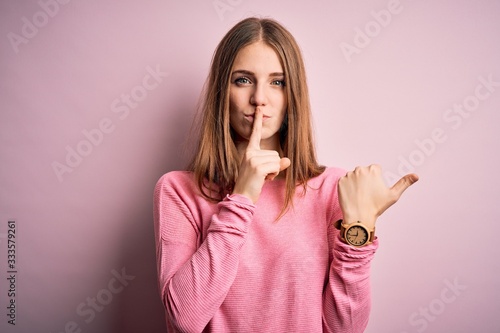 Young beautiful redhead woman wearing casual sweater over isolated pink background asking to be quiet with finger on lips pointing with hand to the side. Silence and secret concept.