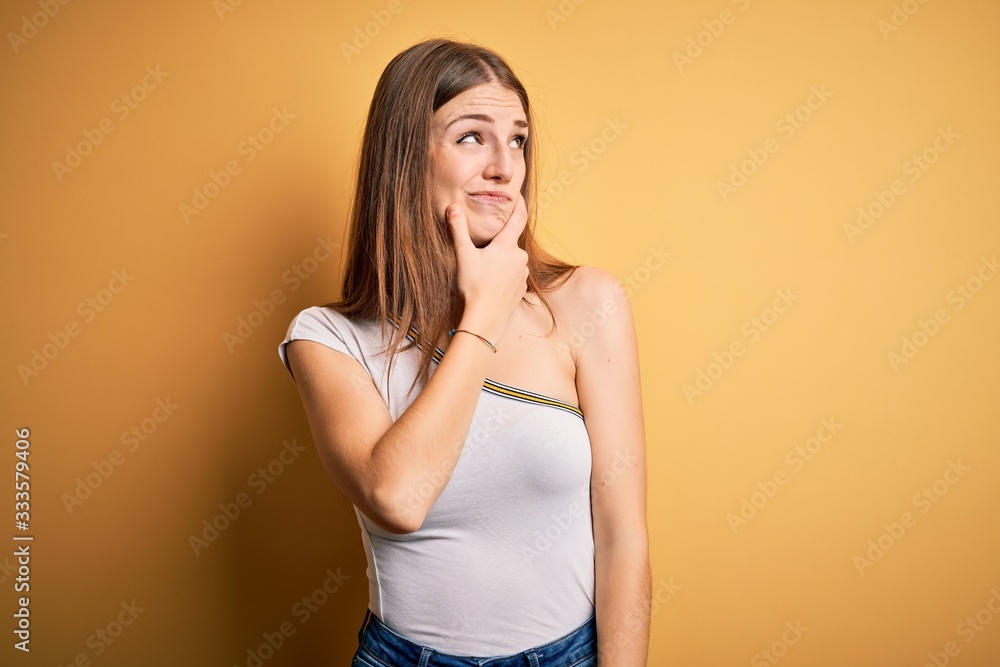 Young beautiful redhead woman wearing casual t-shirt over isolated yellow background Thinking worried about a question, concerned and nervous with hand on chin Young beautiful redhead woman wearing casual t-shirt over isolated yellow background Thinking worried about a question, concerned and nervous with hand on chin