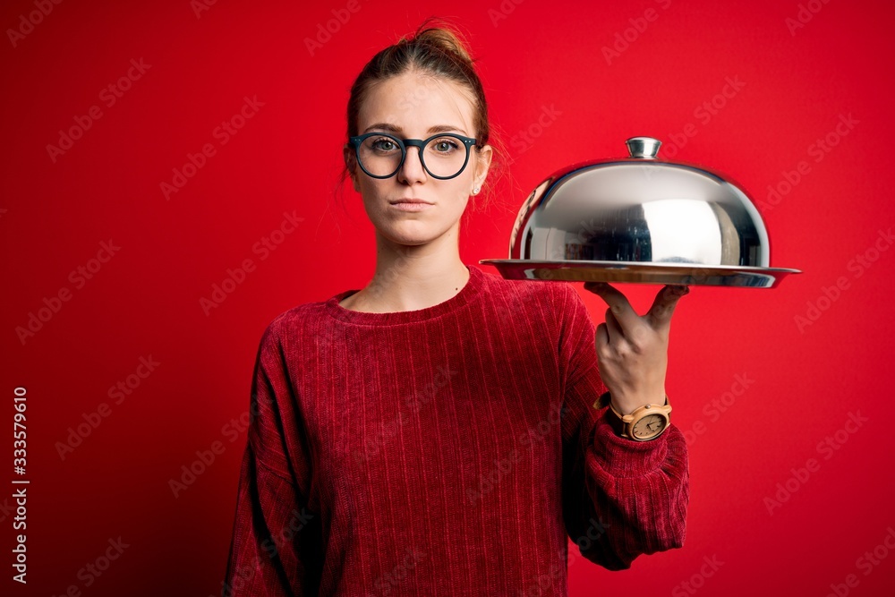Young beautiful redhead woman holding waitress tray over isolated red background with a confident expression on smart face thinking serious Young beautiful redhead woman holding waitress tray over isolated red background with a confident expression on smart face thinking serious