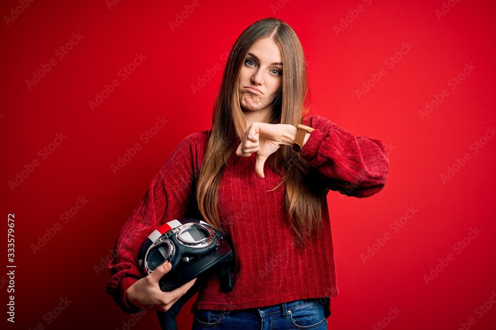 Young beautiful redhead motocyclist woman holding moto helmet over red background with angry face, negative sign showing dislike with thumbs down, rejection concept Young beautiful redhead motocyclist woman holding moto helmet over red background with angry face, negative sign showing dislike with thumbs down, rejection concept