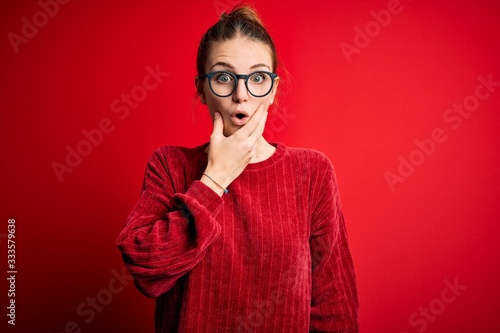 Young beautiful redhead woman wearing casual sweater over isolated red background Looking fascinated with disbelief, surprise and amazed expression with hands on chin