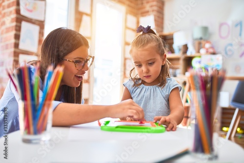Caucasian girl kid playing and learning at playschool with female teacher. Mother and daughter at playroom around toys drawing on magnetic blackboard photo