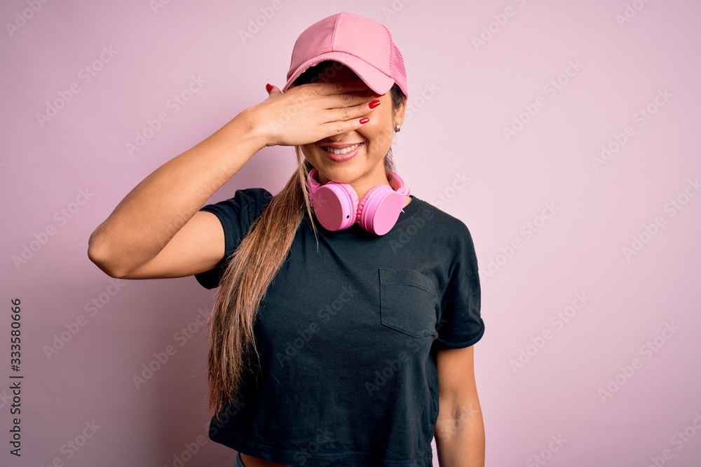 Young beautiful fitness sports woman wearing training cap and headphones over pink background smiling and laughing with hand on face covering eyes for surprise. Blind concept. Young beautiful fitness sports woman wearing training cap and headphones over pink background smiling and laughing with hand on face covering eyes for surprise. Blind concept.