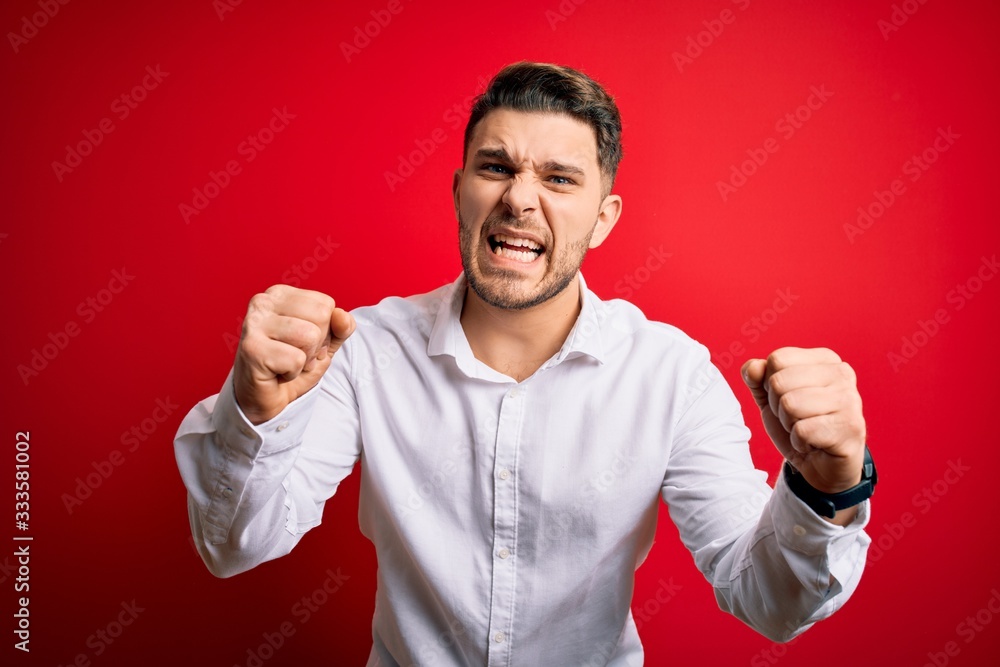 Young business man with blue eyes wearing elegant shirt standing over red isolated background angry and mad raising fists frustrated and furious while shouting with anger. Rage and aggressive concept. Young business man with blue eyes wearing elegant shirt standing over red isolated background angry and mad raising fists frustrated and furious while shouting with anger. Rage and aggressive concept.