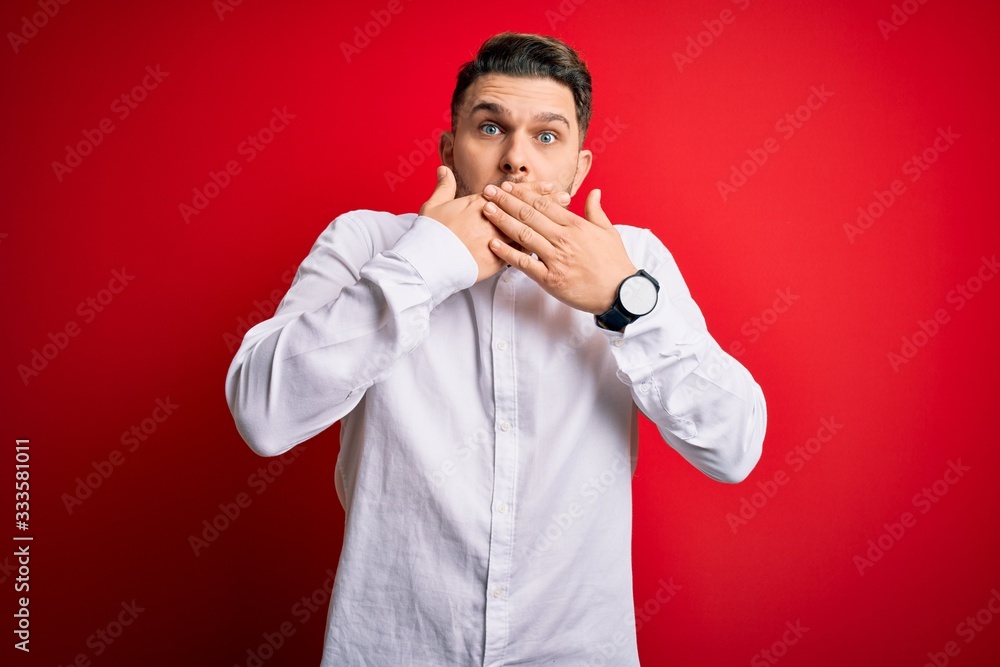 Young business man with blue eyes wearing elegant shirt standing over red isolated background shocked covering mouth with hands for mistake. Secret concept. Young business man with blue eyes wearing elegant shirt standing over red isolated background shocked covering mouth with hands for mistake. Secret concept.