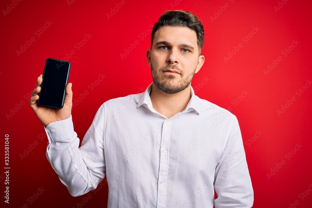 Young business man with blue eyes showing smartphone screen over red background with a confident expression on smart face thinking serious Young business man with blue eyes showing smartphone screen over red background with a confident expression on smart face thinking serious