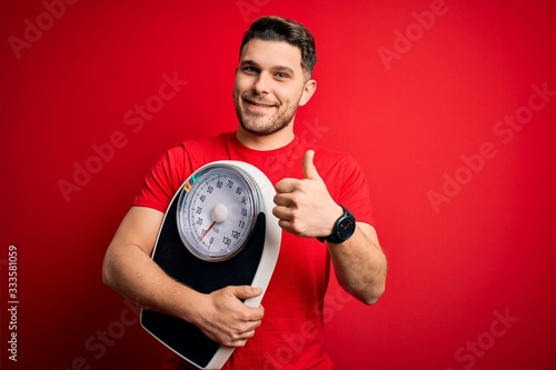 Young fitness man with blue eyes holding scale dieting for healthy weight over red background happy with big smile doing ok sign, thumb up with fingers, excellent sign