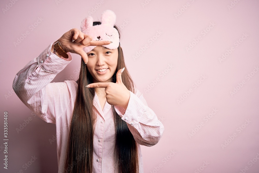 Young asian woman wearing pajama and sleep mask over pink isolated background smiling making frame with hands and fingers with happy face. Creativity and photography concept.