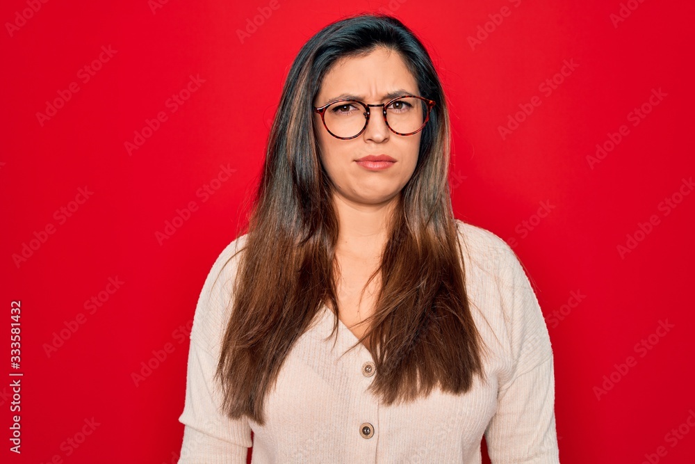 Young hispanic smart woman wearing glasses standing over red isolated background skeptic and nervous, frowning upset because of problem. Negative person.