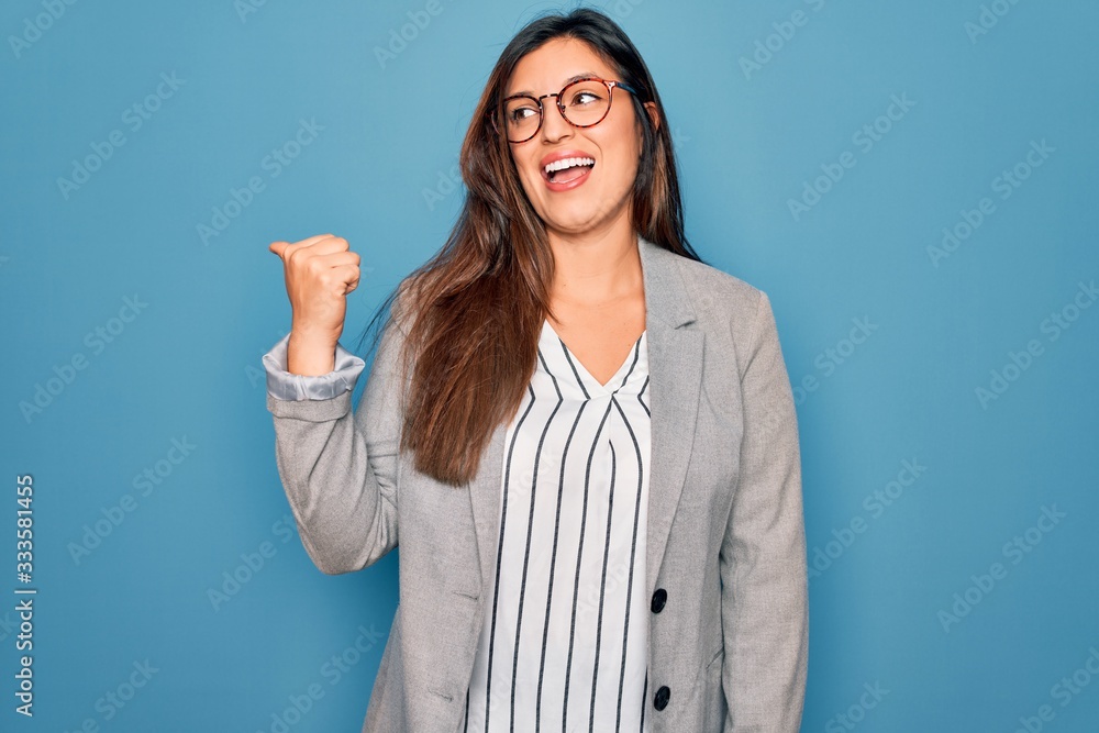 Young hispanic business woman wearing glasses standing over blue isolated background smiling with happy face looking and pointing to the side with thumb up. Young hispanic business woman wearing glasses standing over blue isolated background smiling with happy face looking and pointing to the side with thumb up.