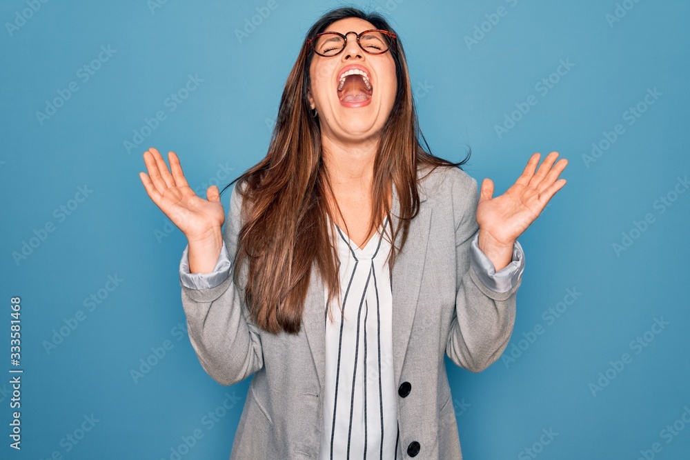 Young hispanic business woman wearing glasses standing over blue isolated background celebrating mad and crazy for success with arms raised and closed eyes screaming excited. Winner concept Young hispanic business woman wearing glasses standing over blue isolated background celebrating mad and crazy for success with arms raised and closed eyes screaming excited. Winner concept