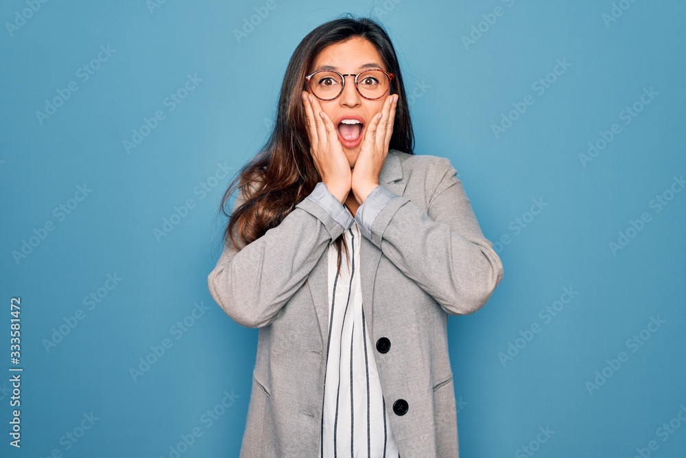 Young hispanic business woman wearing glasses standing over blue isolated background afraid and shocked, surprise and amazed expression with hands on face Young hispanic business woman wearing glasses standing over blue isolated background afraid and shocked, surprise and amazed expression with hands on face