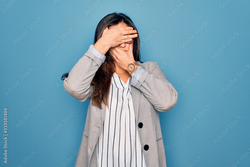 Young hispanic business woman wearing glasses standing over blue isolated background Covering eyes and mouth with hands, surprised and shocked. Hiding emotion Young hispanic business woman wearing glasses standing over blue isolated background Covering eyes and mouth with hands, surprised and shocked. Hiding emotion